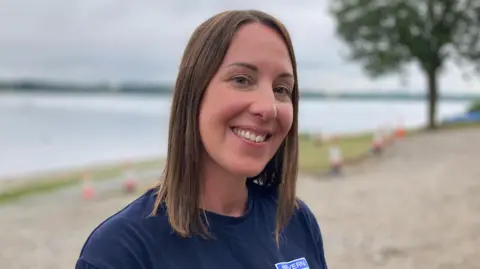 A woman smiles toward the camera. She has shoulder length brown hair and is wearing a dark blue t-shirt with a Severn Trent Water logo on it. There is a lake behind her.