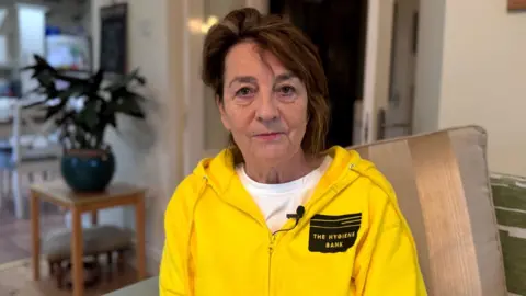 Eileen Brown with short brown hair and is pictured staring at the camera as she wears a yellow zip-up jumper with 'The Hygiene Bank' written on it. She is sitting on a sofa and there is a table with a plant pot on it in the background.