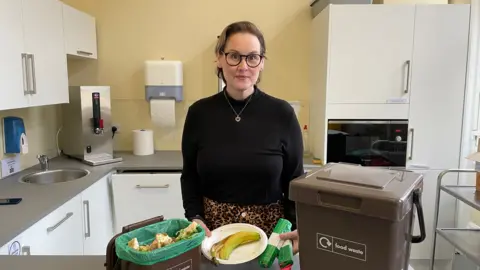 Mark Ansell/BBC Councillor Danielle Durrans, Cabinet Member for Public Services and Communities at Calderdale Council, in the Halifax Town Hall staff kitchen. She is wearing a black jumper and glasses and is behind a brown caddy with food waste inside and a bigger brown big which is where the bags of rubbish are placed for collection. 