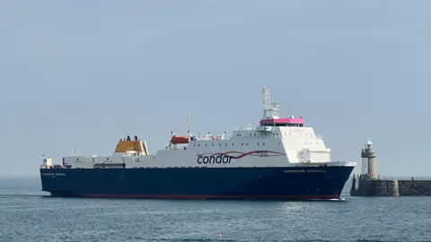 A side view of a cargo ship. The vessel is large and is blue and white. It is anchored on a calm sea. The sky is blue. The Condor logo is printed down the side of the boat.