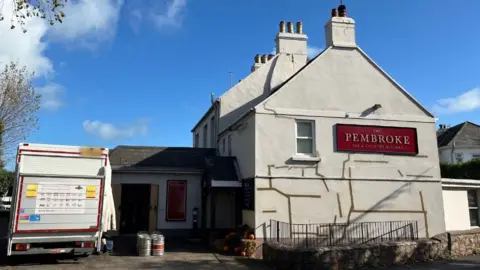 The Pembroke pub in Jersey. The building has white walls and a red sign with Pembroke written in gold letters. A lorry is outside with kegs of beer being delivered to the pub.