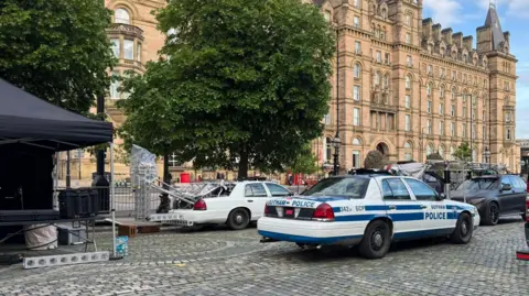 @EGGBOY_03 A blue and white liveried Gotham police car is on St George's Plaza surrounded by other vehicles and a filming tent, with a tree and grand old building behind.
