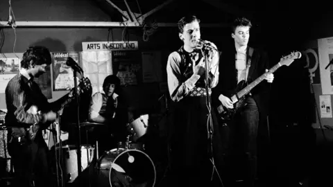 The Dreamboys/Harry Papadopoulos/Street Level Photoworks A black and white image of The Dreamboys performing on stage during the late 70s or early 80s. Peter Capaldi is singing at a microphone and there are two guitarists and a drummer