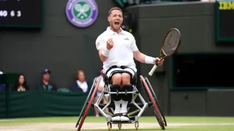 PA Media Alfie Hewett, wearing white tennis gear, pumps his right fist in triumph, on the court at Wimbledon. He is in a specialist wheelchair and is holding a racket in his left hand.