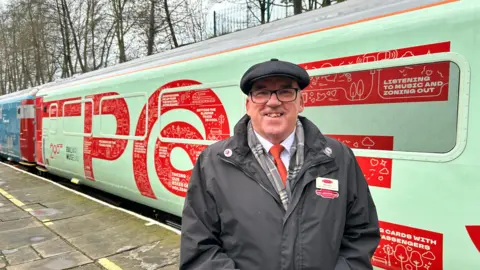 Network Rail A man wearing a grey coat and dark grey flat cap is smiling while standing in front of a pistachio green train. It has writing on it including 'listening to music and zoning out' and 'railway museum'.