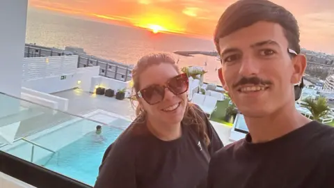 Caprice Betts and her partner Arron Sanchez Garcia standing on a balcony with a swimming pool beneath them and a view out to sea and a setting sun. She is on the left wearing a black T-shirt, has dark brown hair pulled back in a pony tail and is wearing dark glasses. He is on the right with black short hair, a black moustache and is wearing a black T-shirt. They are both smiling. 