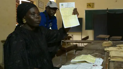 A woman in a black top displays a ballot paper. She is sitting alongside a man wearing blue, at a desk with many ballot papers on top inside a voting centre on Dakar, Senegal. 
