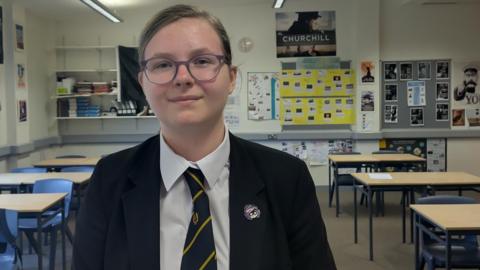 Emily is standing in a classroom looking into the camera with a slight smile. She is wearing a school uniform 