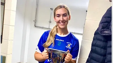 Aislín Ní Choinn A woman with blonde hair is looking at the camera. She is wearing a blue GAA jersey, and is holding a silver trophy. She is in a changing room.