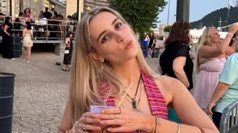 Mia Harrison. She has long blonde hair and is wearing a pink striped top and several necklaces. She is pictured tilting her head to the side, pouting and looking at the camera. She is holding a drink by her chest. 