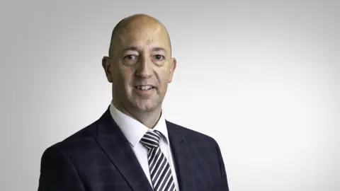Paul Vane pictured in a black check suit. Black and white stripey tie and white shirt. He is smiling. He is middle aged with a bald head. He's being photographed in a studio, with a blank background.