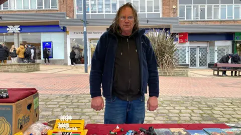 A man wearing a dark blue fleece over a black fleece standing behind a market stall table.