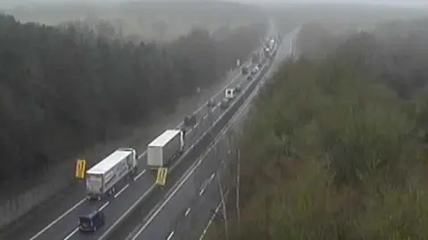 The M26 seen from above with lorries and cars travelling on one side of the road and an empty carriageway on the other side. There are trees either side.