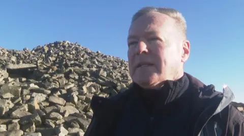 A man with short grey hair, wearing a navy coat. He is standing in front of a pile of rocks and there is a blue sky above. 