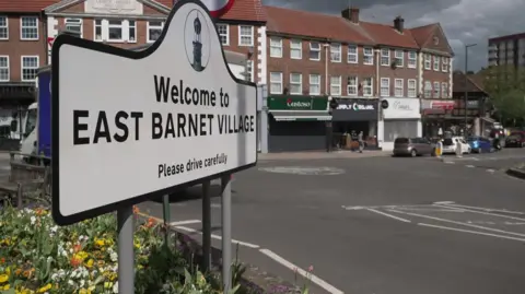 A sign that says Welcome to East Barnet Village stands in by the side of a road. There are shops in the background and a flowerbed in the foreground. 