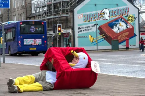 Nuart 2026 Man in a seagull costume, lying on the pavement, with an artwork of a seagull in the background on a wall that he is recreating.