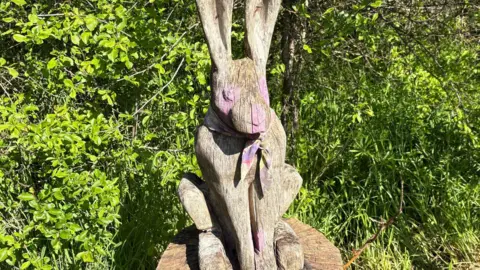 A wooden hare with pink paint splashed on its eyes, nose and paws.