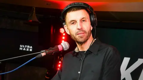 Getty Images Tim Westwood, with a black shirt on, wearing headphones with a microphone in front of him - there are red studio lights behind him and a black backdrop with the words "ultra haze" 