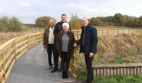 Three men and a woman pictured standing on a fenced boardwalk with trees in the background. They are all looking towards the camera and smiling.