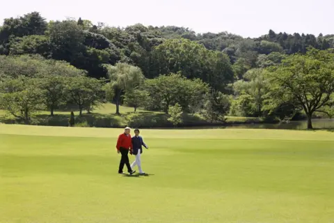 Getty Images Donald Trump i Shinzo Abe chodzą po zielonym, otwartym fairway na środku odległości z dużą ilością drzew za nimi w oddali. Trump ma na sobie czerwoną kurtkę i ciemne spodnie; Abe ciemną kurtkę i białe spodnie. Wydają się głęboko w rozmowie 