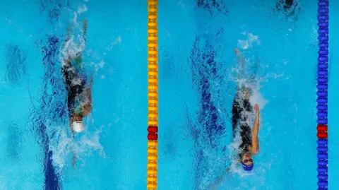 Reuters A pool is seen from the top with two people swimming in lanes. There is a yellow lane marker to the right of one swimmer and a blue marker to the right of the other swimmer. Both swimmers are doing fast backstroke and both are wearing swimming caps.