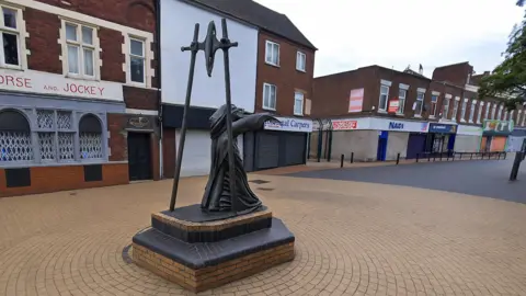 General view of Bilston's high street. A black statue of a woman bent over carrying a bag on her back in front of a wheel supported by two sticks. In the background is an empty high street