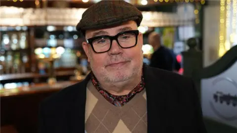 Cliff Parisi in a tartan hat and black-framed glasses standing in front of a bar in a pub. He has stubble and is wearing a flat-cap. He appears to be wearing a black coat or suit jacket.