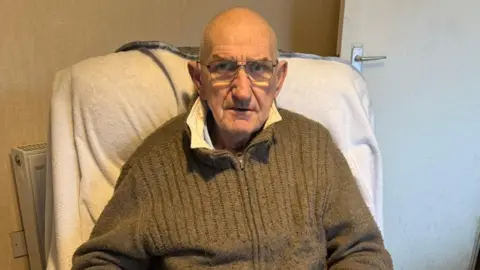 John Hargreaves, who is bald, wears glasses and is wearing a light brown zip-up fleece, sits in an arm-chair looking into the camera with a serious expression. 