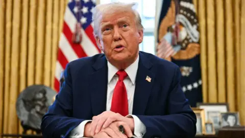 Donald Trump photographed behind a desk in the Oval Office with gold curtains and US flags behind him. He is visible from the waist up, with his arms crossed on a desk in front of him. He is wearing his customary navy suit and bright red tie. 