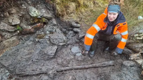 A female archaeologist wearing an orange high visibility jacket crouches down next to mud-covered stones and a timber post.