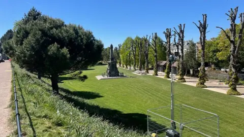 Canterbury City Council A wide view of Dane John Gardens shows trimmed lawns, a memorial, rows of pollarded trees and temporary fencing under blue sky.