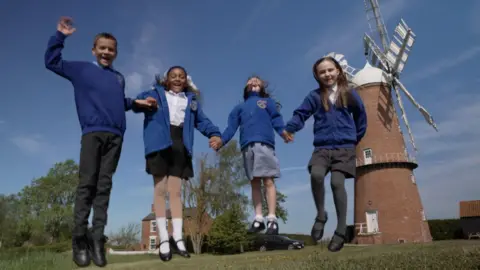 Primary school pupils wearing blue jumpers with windmill logos smile at the camera while jumping in front of the newly repaired mill