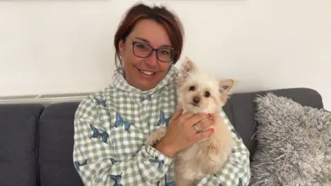 Anabel Sharma Anabel Sharma sitting on a grey sofa, wearing a light green gingham hoody with printed blue butterflies. She is smiling while carrying a puppy which is looking at the camera.