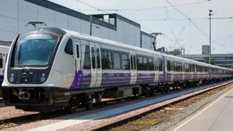 TfL An Elizabeth Line train is seen pulling into a train station. The train has purple colouring on its bottom half and large glass office buildings can be seen in the background
