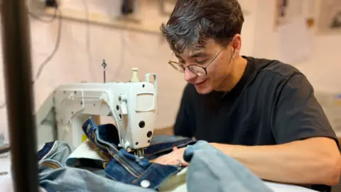 Cosmin Diaconu sits at a sewing machine repairing a pair of denim jeans. He is wearing a black T-shirt and has glasses on and black hair. 