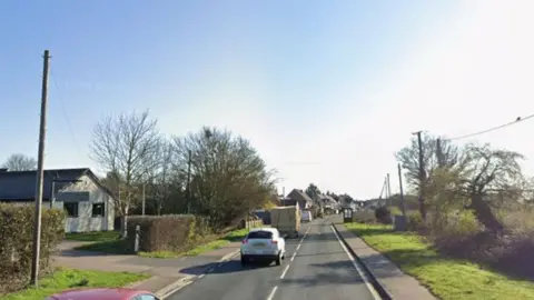 A view of Stortford Road, with cars travelling away from the camera, and trees and bushes either side. It is sunny with a blue sky above.