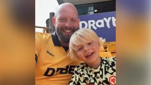 Tom, who has a beard, is wearing a Wolverhampton Wanders FC shirt and smiling next to son Rory, who has blond hair. They are both smiling at the camera while sitting in the stadium's stand.