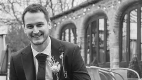 Thames Valley Police/Family Jason Wallis: A black-and-white image of Jason Wallis wearing a dark jacket and tie, with a flower in the right lapel, and a white shirt. . He is in front of a building with arched windows. A man and woman can be seen behind Wallis's right shoulder. 
