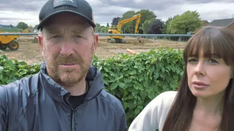 A man with a beard wearing a grey zip-up jacket and a black baseball cap. He is stood next to a woman with long brown hair and a fringe. They both have a serious expression. They are stood in their garden which has a construction site behind it with two yellow diggers on the site. 