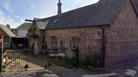 A google map image of the former blacksmith's forge on Lower Road, Eastbourne, which is slated for redevelopment. The red-brick building has green window frames and a gate entry. 