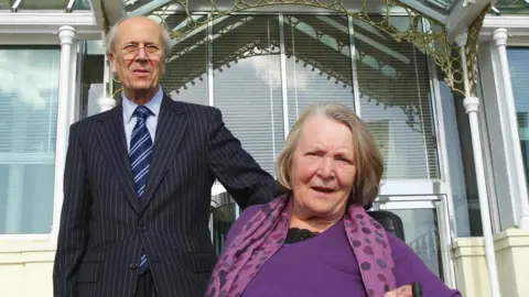 PA Media A man in a pinstriped suit, a blue striped tie and glasses standing behind a woman in a wheelchair. She is wearing a purple top and a purple scarf. Behind them is a cream building with windows.