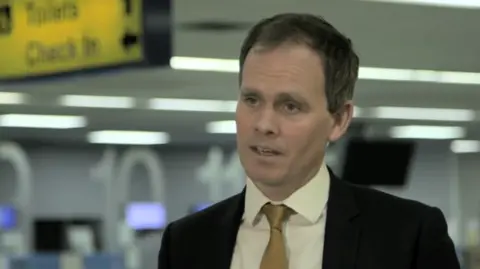 Andrew Bell, chief executive of Regional & City Airports, the operator of Bournemouth Airport, giving an interview at what looks like the inside of an airport. There is a yellow sign for toilets and check in on the ceiling above him.
