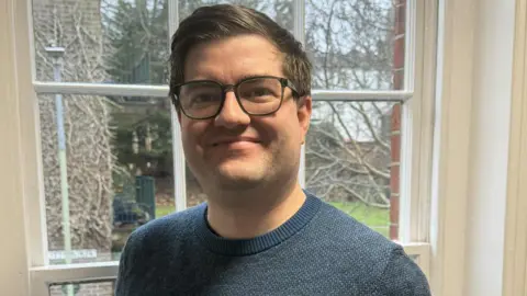 A man standing in front of a window with a garden in the background. He has brown hair and black glasses and is wearing a black jumper
