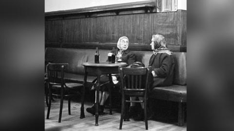 Two older women wearing headscarves sit at a table in an pub in 1977, both having a bottle and glass of stout in front of them