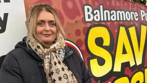 Joanne Skinner is looking into the camera. She's wearing a brown scarf with star patterns on it and a padded black coat. A large sign saying save our school is behind her.