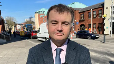 Gary Ridley faces the camera. He is standing in front of a road and is wearing a grey suit jacket and grey tie.