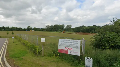 Google Maps A white and red sign reads SOLD and is pitched against a metal fence. Behind the sign and fence is a large green field with one yellow digger moving piles of dirt. 