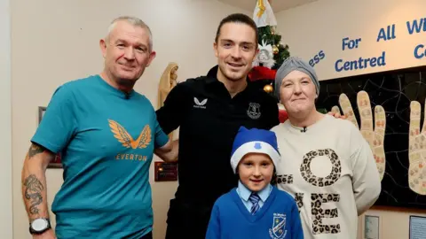EFC Group shot of Grealish with his arms around mum Gemma, wearing a grey turban cap and white jumper with the word Love, and grandad Keith in a blue Everton shirt on the left. Georgie in his blue Everton santa hat and blue school uniform stands in front of them. 