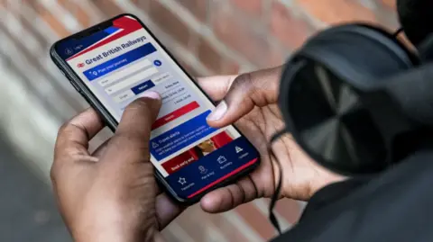 Department for Transport The Great British Railways app being used on someone's phone - it can be seen being held in their hands with the GBR app visible on the screen - an obscured brick work is visible in the near ground