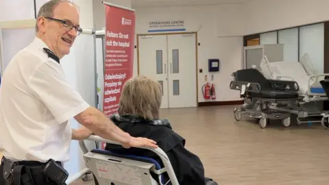 Health and Care Jersey Dressed in a shirt with epaulettes and black trousers, a smiling man is pushing a patient in a wheelchair who has their back to us. He is wearing glasses and has short cropped dark hair. They are in a corridor with several hospital beds.